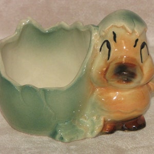 May include: A ceramic planter shaped like a cartoon animal peeking out of a cracked egg. The planter is glazed in shades of green and brown, with painted details on the animal's face.