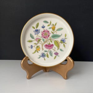 May include: A small, round, white ceramic plate with a gold rim, decorated with a floral pattern in pink, blue, yellow, and green. The plate is presented on a gold-coloured stand, against a black and white backdrop.