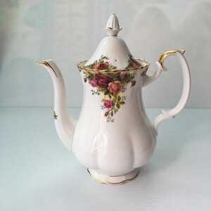 Op de afbeelding: Witte porseleinen theepot met een gouden rand en een bloemenpatroon van rozen en bladeren. De theepot heeft een deksel met een knop bovenop.