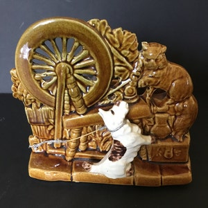 May include: A ceramic figurine of a dog and cat with a spinning wheel. The dog is white with brown spots and is looking at the spinning wheel. The cat is brown and is sitting on a ledge. The spinning wheel is brown and has a white string attached to it. The figurine is made in a brown glaze.