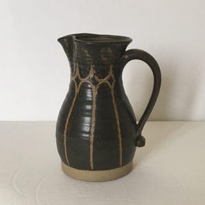 Könnte beinhalten: Ein brauner Keramik Krug mit einer hellbraunen Glasur und einem geometrischen Muster. Der Krug hat einen breiten Boden und einen schmalen Hals.