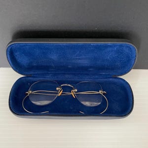 May include: Rimless spectacles with gold wire frames and clear lenses are displayed inside an open, blue velvet-lined case. The case is black on the outside. The glasses have a classic, vintage aesthetic.