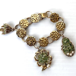 May include: Vintage gold-coloured bracelet with textured, circular links. The bracelet has three charms: a cluster of green stones in a flower shape, a teardrop charm with green stones and pearls, and a fan-shaped charm with green stones.