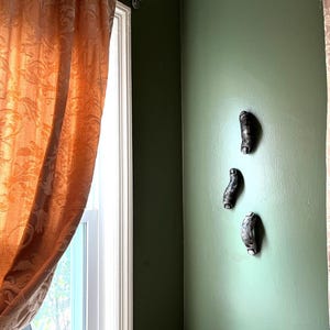 May include: Three small, curved, dark gray and black textured wall-mounted sculptures are arranged vertically on a solid olive green wall. Each piece has a smooth, rounded shape with subtle variations in tone and a slightly weathered appearance.