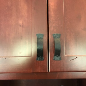 May include: Two black metal cabinet door pulls with a simple, curved design. The pulls are mounted on a dark wood cabinet door.