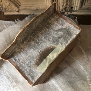 May include: A vintage cardboard box with a worn, faded brown exterior. The box has a slightly curved top and a piece of tape on the side.