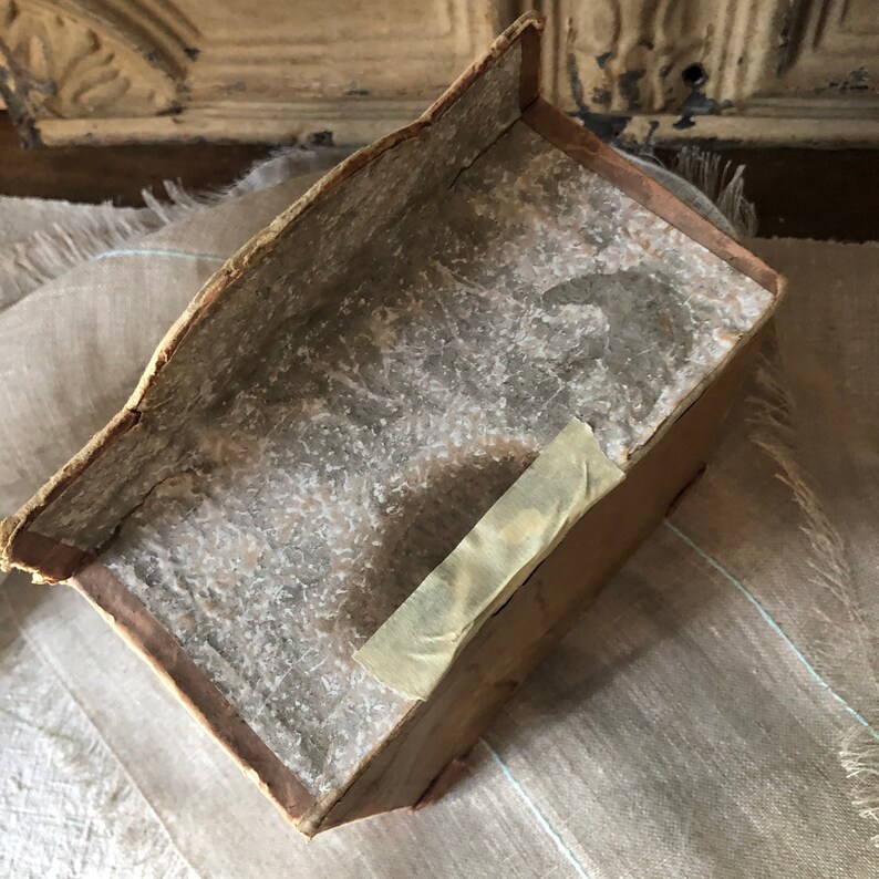 May include: A vintage cardboard box with a worn, faded brown exterior. The box has a slightly curved top and a piece of tape on the side.