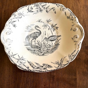 May include: Cream-coloured, scalloped-edge serving dish with a black and white bird and floral design. The centre features two birds in a marshy landscape. The rim is decorated with floral and scroll patterns. A decorative piece for the home.