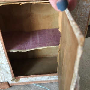 May include: A small cardboard cabinet with a single door that opens to reveal two shelves. The shelves are lined with purple paper. The cabinet is made of cardboard and has a white painted finish.