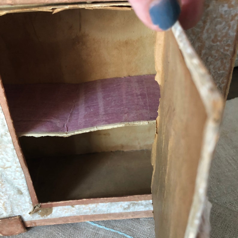 May include: A small cardboard cabinet with a single door that opens to reveal two shelves. The shelves are lined with purple paper. The cabinet is made of cardboard and has a white painted finish.