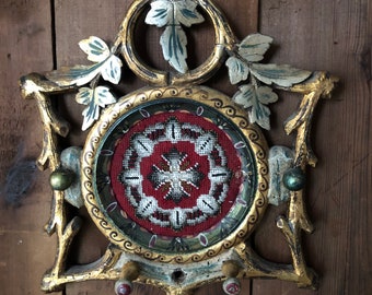 May include: A gold-painted wooden frame with intricate carvings of leaves and vines. A round, beaded design is displayed in the center of the frame.