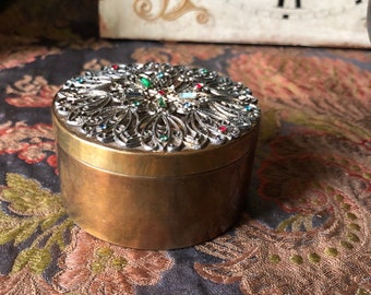 May include: A round brass trinket box with a filigree lid decorated with colorful rhinestones. The lid features a floral design with a central starburst.
