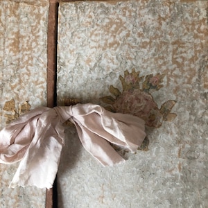 May include: Two antique books bound in brown paper with a delicate pink ribbon tied around them. The covers are decorated with a floral design in shades of brown and gold.