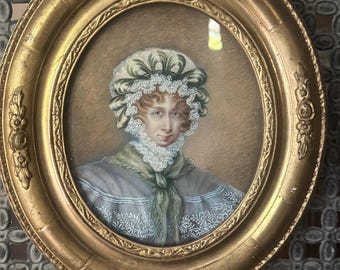 May include: An oval portrait of a woman in a gold frame. The woman is wearing a bonnet with green and white trim, a lace collar, and a blue shawl. The frame is ornate with a gold finish.