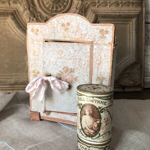 May include: A vintage, white and pink paper box with a pink ribbon and a round, white and gold tin with the text "Nell Gwynne" and a woman in a dress holding a bird.