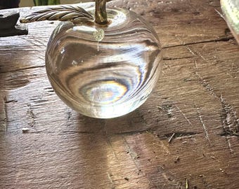 Könnte beinhalten: Ein klarer Glas-Apfel-Briefbeschwerer mit goldfarbenem Stiel und Blatt. Das Glas hat ein Wirbelmuster, und der Apfel liegt auf einer rustikalen Holzoberfläche. Stiel und Blatt sind detailliert.