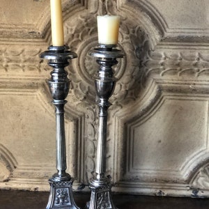 May include: Two antique silver candlesticks with lit candles. The candlesticks have ornate bases and detailed designs. One candlestick holds a tall, thin candle, while the other holds a shorter, wider candle. The background is a weathered, light-colored wooden door.