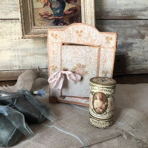 May include: A vintage, ornate, wooden frame with a pink and white floral design. The frame is open and has a white ribbon tied around it. A vintage, cylindrical tin container with a gold lid and a floral design is in front of the frame. The tin container has the text "Nell Gwynne" and "Yardley" printed on it.