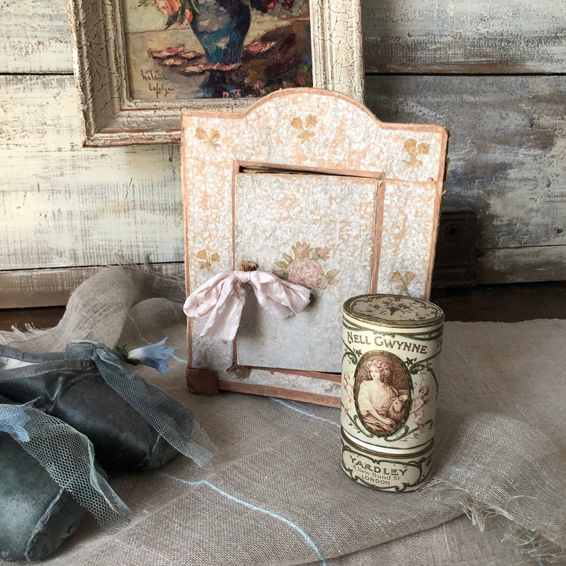 May include: A vintage, ornate, wooden frame with a pink and white floral design. The frame is open and has a white ribbon tied around it. A vintage, cylindrical tin container with a gold lid and a floral design is in front of the frame. The tin container has the text "Nell Gwynne" and "Yardley" printed on it.