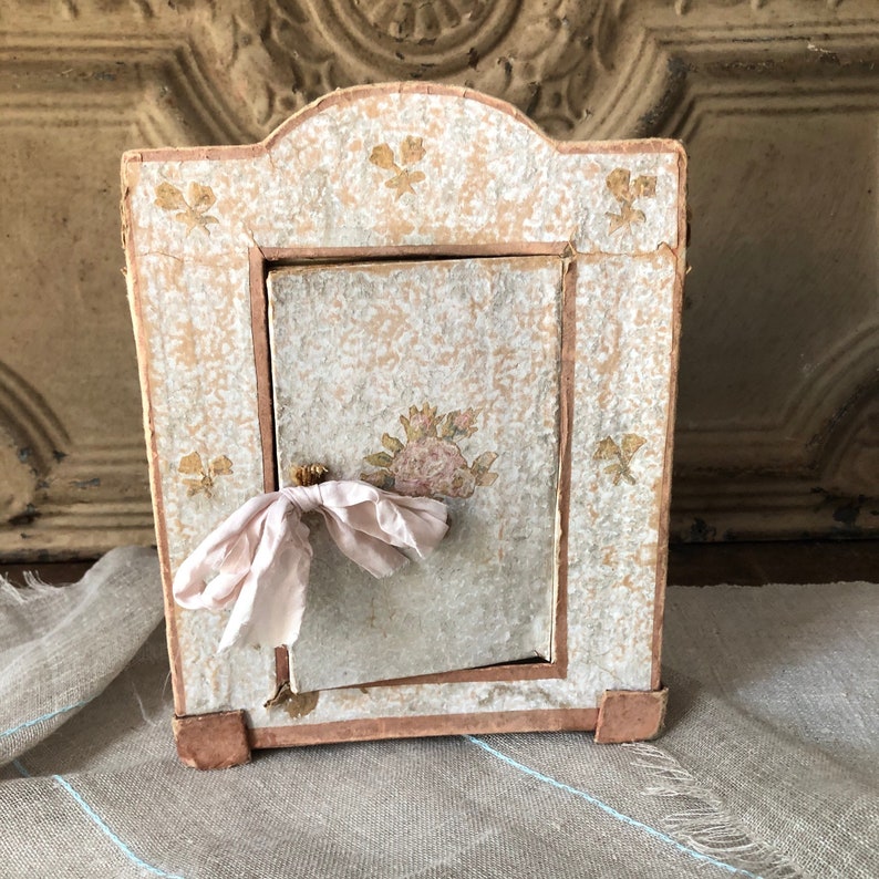 May include: A small, white, wooden box with a floral design and a pink ribbon tied around the handle. The box has a hinged door and is decorated with a floral pattern.