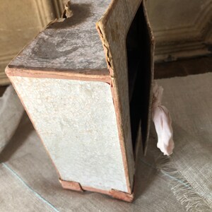 May include: A small, vintage cardboard dollhouse with a white exterior and brown trim. The dollhouse has a pink fabric hanging from the side.