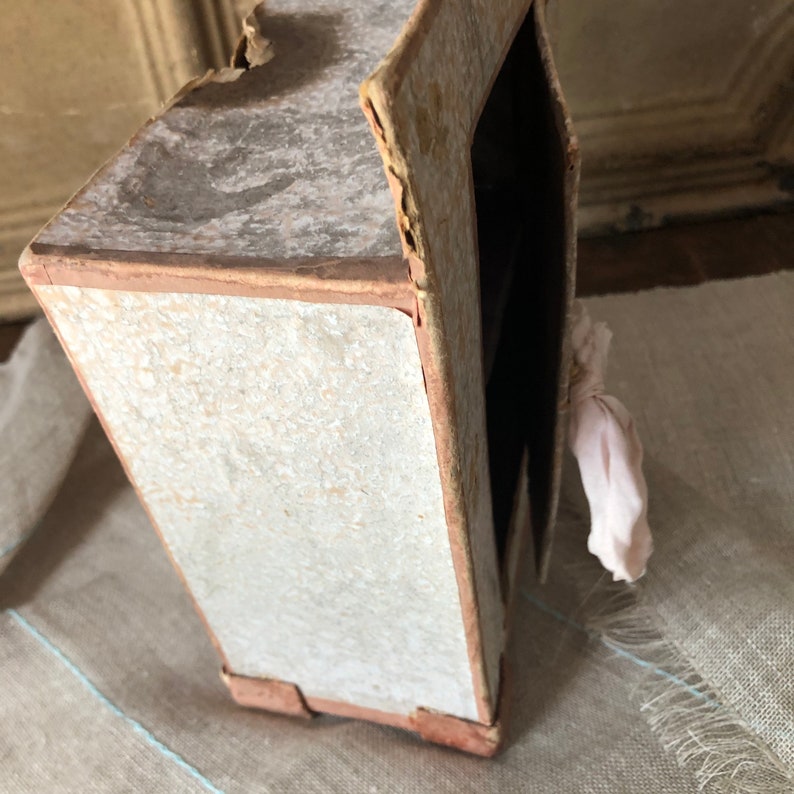 May include: A small, vintage cardboard dollhouse with a white exterior and brown trim. The dollhouse has a pink fabric hanging from the side.