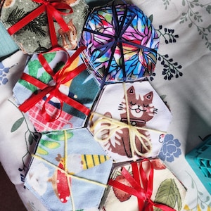 Puede incluir: Un conjunto de seis almohadillas de desmaquillaje reutilizables de forma hexagonal. Las almohadillas están hechas de tela suave y tienen diferentes patrones coloridos, incluyendo un gato, fresas y champiñones. Las almohadillas están atadas juntas con una cinta roja.