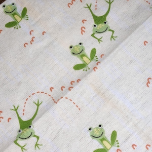May include: White fabric with a repeating pattern of green frogs jumping. The frogs are outlined in black and have a white smile. The frogs are jumping over a dotted line of orange footprints.