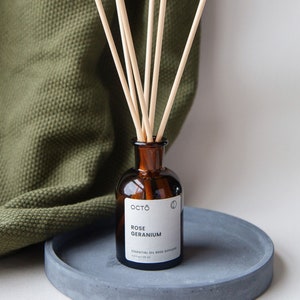 Op de afbeelding: Een bruine glazen fles met een etiket met de tekst "OCTO ROSE GERANIUM ESSENTIAL OIL REED DIFFUSER 4.2 fl oz / 125 ml" en een set houten diffuserstokjes.
