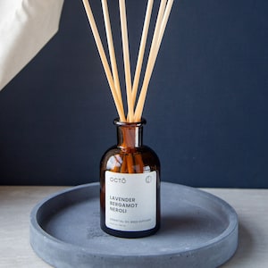 May include: A brown glass bottle with a label that reads "OCTO Lavender Bergamot Neroli Essential Oil Reed Diffuser 125 ml". The bottle is filled with a clear liquid and has wooden sticks sticking out of the top. The bottle is sitting on a grey concrete tray.