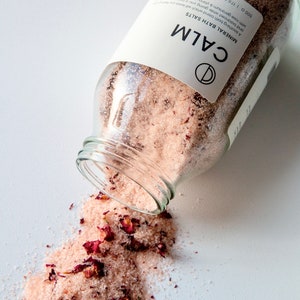 Pode incluir: Um frasco de vidro de sais de banho minerais cor-de-rosa com pétalas de rosa secas, etiquetado como "CALM MINERAL BATH SALTS" e "500 g | 17.6 OZ e". O frasco está tombado, derramando os sais sobre uma superfície branca.