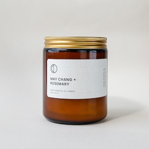 May include: A brown glass jar candle with a gold lid and a white label. The label reads "MAY CHANG + ROSEMARY" and "PURE ESSENTIAL OIL CANDLE". The candle is 250 ml. The text "HAND POURED IN LONDON BY OCID" is also visible.
