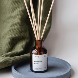 Può includere: Un flacone di vetro marrone con un'etichetta bianca su cui è scritto "OCTO" e "MORNING LIGHT ESSENTIAL OIL REED DIFFUSER 6.7 fl oz / 200 ml". Il flacone è riempito con un liquido trasparente e ha bastoncini di legno inseriti al suo interno. Il flacone è appoggiato su un sottobicchiere di cemento grigio.