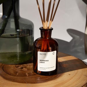 Puede incluir: Una botella de vidrio marrón con una tapa de madera y una etiqueta que dice "OCTO MORNING LIGHT ESSENTIAL OIL REED DIFFUSER 4.2 fl oz (125 ml)". La botella está llena de un líquido transparente y tiene palitos de madera que sobresalen.