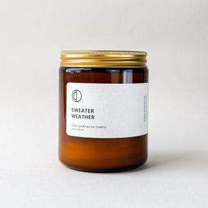 Peut inclure: Une bougie en pot de verre brun avec un couvercle doré. L'étiquette indique "Sweater Weather" et "Pure Essential Oil Candle". La bougie est de 250 ml. Le pot est sur un fond neutre.