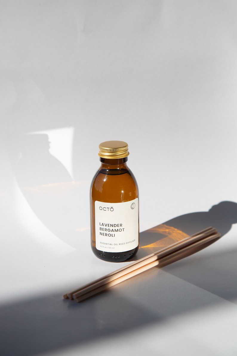 May include: A brown glass bottle with a gold lid and a white label that reads "Octo Lavender Bergamot Neroli Essential Oil Reed Diffuser 4.2 fl oz / 120 ml". The bottle is sitting on a white surface with a set of wooden diffuser sticks.