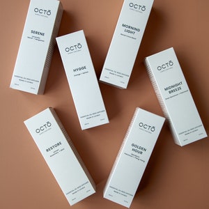 May include: Six white essential oil reed diffusers with black text on a light brown surface. The diffusers are labelled with different scents: Serene, Morning Light, Hygge, Midnight Breeze, Restore, and Golden Hour.