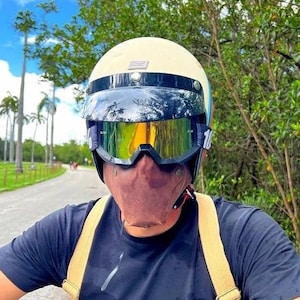 Pode incluir: Uma pessoa vestindo uma camiseta preta com um logotipo branco e preto que diz "Brothers and Motard Specialists" está usando um capacete bege com uma viseira escura e uma máscara facial marrom. A pessoa está dirigindo uma motocicleta em uma estrada pavimentada com palmeiras de ambos os lados.