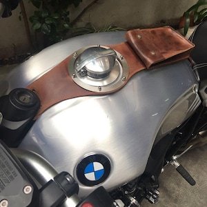May include: A silver motorcycle gas tank with a brown leather strap and a small brown leather pouch. The tank has a round, silver gas cap with a BMW logo on the front.