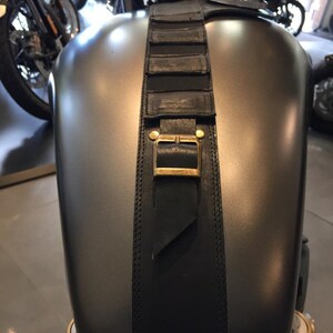 Puede incluir: Una correa de cuero negra con una hebilla de lat&oacute;n est&aacute; unida al tanque de gasolina de una motocicleta. La correa tiene varios compartimentos cosidos.