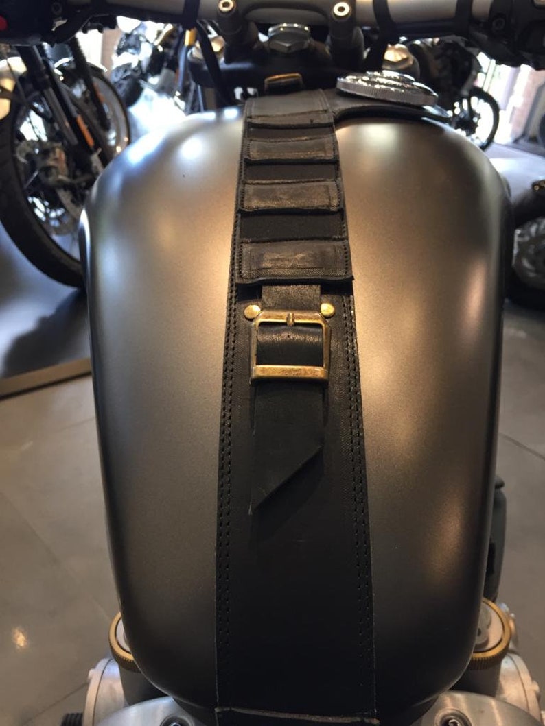 Puede incluir: Una correa de cuero negra con una hebilla de lat&oacute;n est&aacute; unida al tanque de gasolina de una motocicleta. La correa tiene varios compartimentos cosidos.