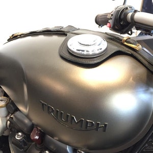 Puede incluir: Un tanque de gasolina de motocicleta gris con una correa de cuero negro y una tapa de gasolina plateada. El tanque tiene la palabra "TRIUMPH" en relieve.