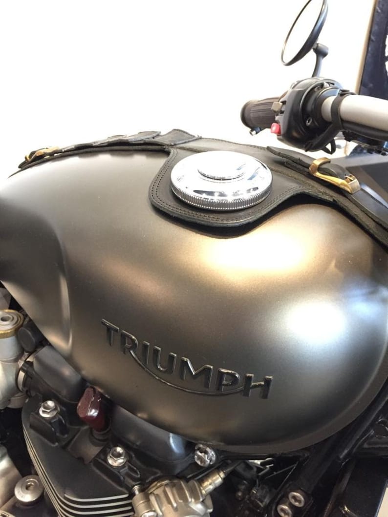 Puede incluir: Un tanque de gasolina de motocicleta gris con una correa de cuero negro y una tapa de gasolina plateada. El tanque tiene la palabra "TRIUMPH" en relieve.