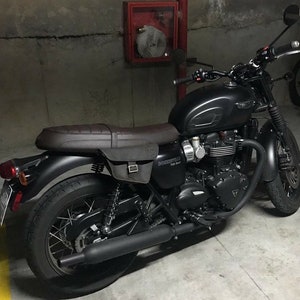 May include: A black Triumph Bonneville T120 motorcycle with a brown leather seat and saddlebags. The motorcycle is parked in a garage with a fire extinguisher in the background.