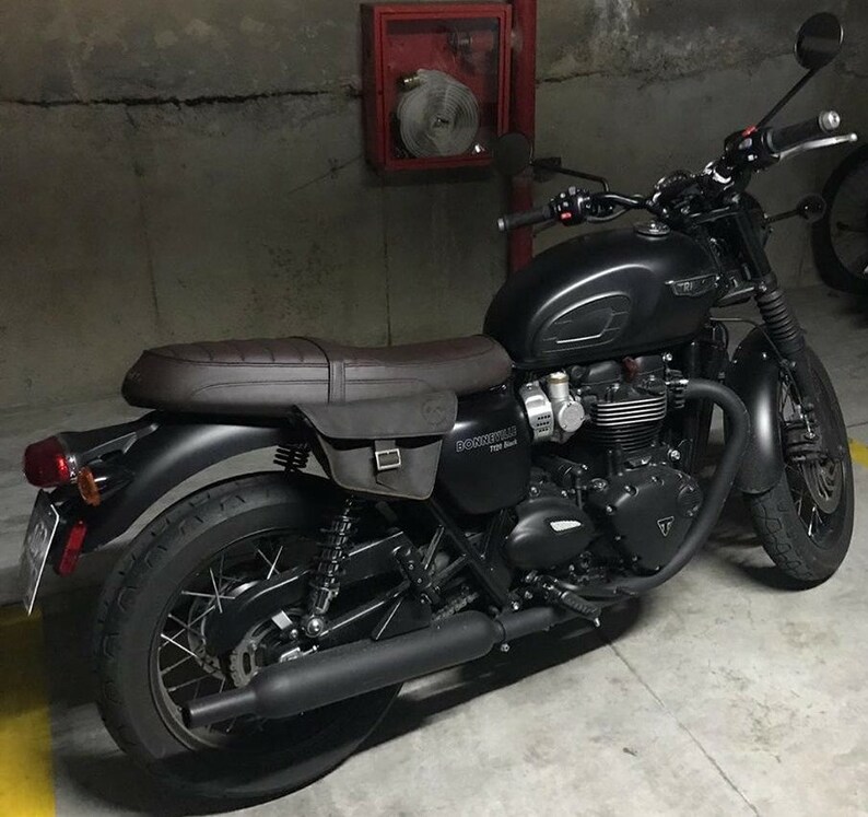 May include: A black Triumph Bonneville T120 motorcycle with a brown leather seat and saddlebags. The motorcycle is parked in a garage with a fire extinguisher in the background.