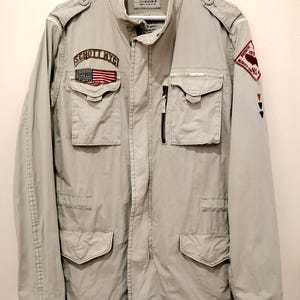 Könnte beinhalten: Hellgrüne Jacke mit mehreren Taschen und Aufnähern. Die Jacke trägt den Schriftzug "SCHOTT NYC" und einen amerikanischen Flaggenaufnäher. Sie hat einen Reißverschluss und Knöpfe. Die Jacke hängt an einem weißen Kleiderbügel.