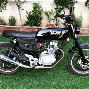 May include: A black motorcycle with a custom seat, a black leather saddlebag, and a black exhaust pipe. The motorcycle has a silver engine and a black frame.