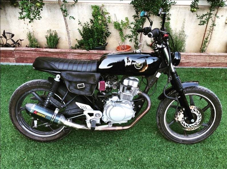 May include: A black motorcycle with a custom seat, a black leather saddlebag, and a black exhaust pipe. The motorcycle has a silver engine and a black frame.