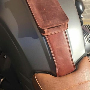 May include: A brown leather motorcycle seat with a brown leather pouch attached to the front. The pouch has a flap with a metal snap closure.