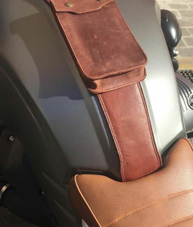 May include: A brown leather motorcycle seat with a brown leather pouch attached to the front. The pouch has a flap with a metal snap closure.